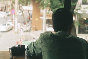 Rear view of a young man with a vintage camera sitting by a cafe window looking at the street